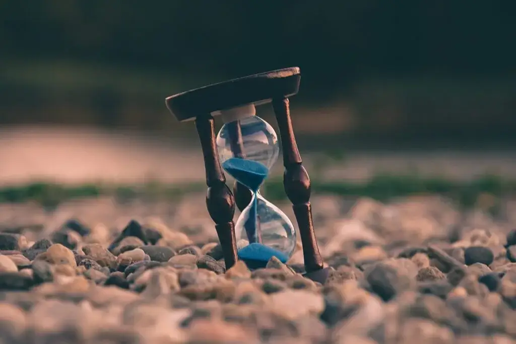 Hourglass with blue sand running through it, resting on small pebbles at dusk