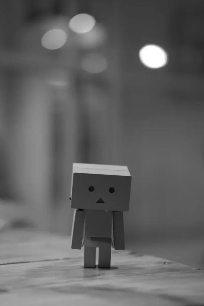 Small cardboard robot figure standing alone on a wooden surface, with blurred lights in the background