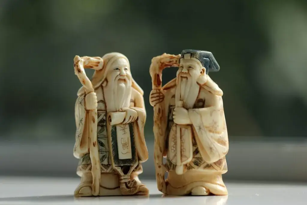 Two carved figurines of long-bearded elders holding staffs, on a light surface against a blurred green background